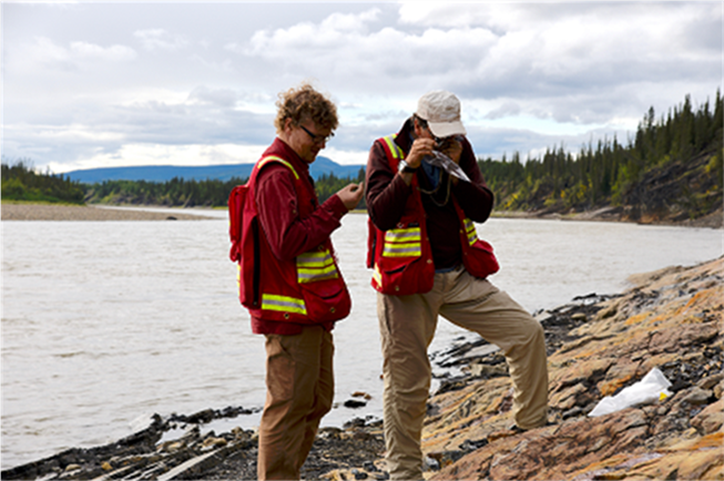A photo of two highly-qualified personnel looking at rock formations on the edge of a lake.