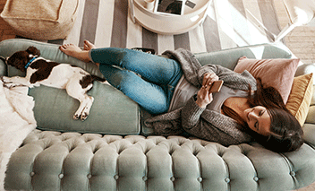 Woman using her cellphone while relaxing on a couch with a dog at home.