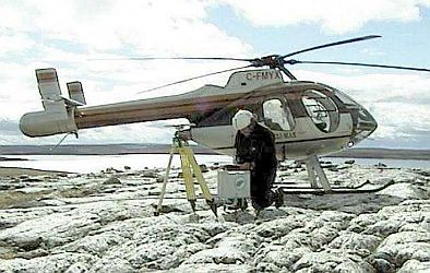Technician kneeling on rock outcrop taking measurements with a CG-5 gravimeter with a helicopter in the background