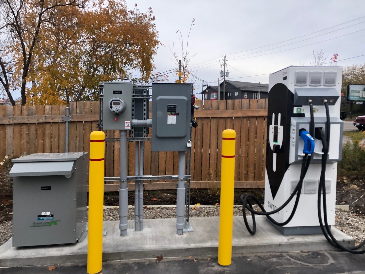 An electric vehicle charger, situated in a parking lot in front of a fence.