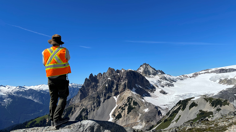 Researcher wearing a safety vest with back to camera looks out at snow capped mountain vista.