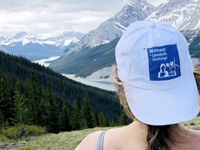 Over-the-shoulder of Amanda looking out at a Rocky Mountain scene. Wearing white cap with text: Women Cleantech Challenge 