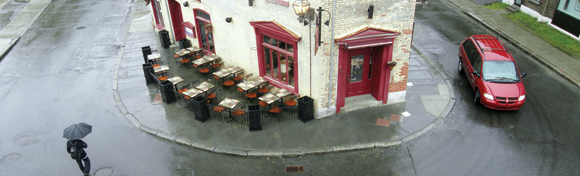 aerial photograph of an old street corner in Quebec City with a man walking on the street in the rain under his open umbrella and empty tables nearby on the sidewalk